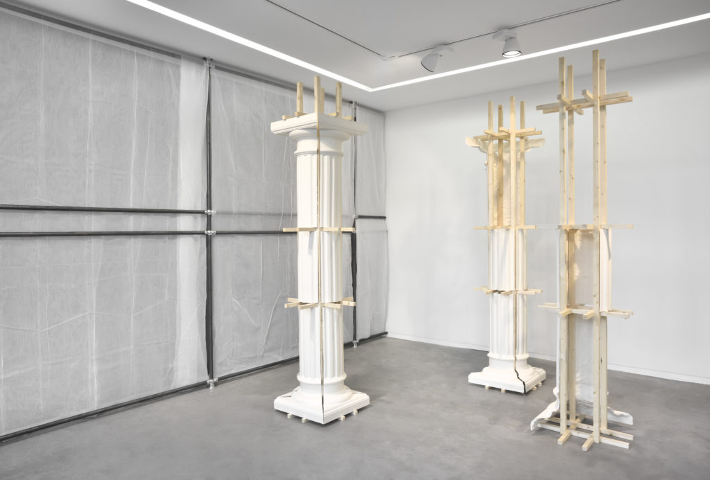 Cyril Zarcone, Colonnes, 2016. Plâtre, bois, 240 x 55 x 55 cm. En arrière-plan, Protection, 2016. Filets de chantier, tubes métalliques. Courtesy Galerie Eric Mouchet. Vue de l’exposition personnelle « Re/productions », 2016, Galerie Eric Mouchet, Paris.